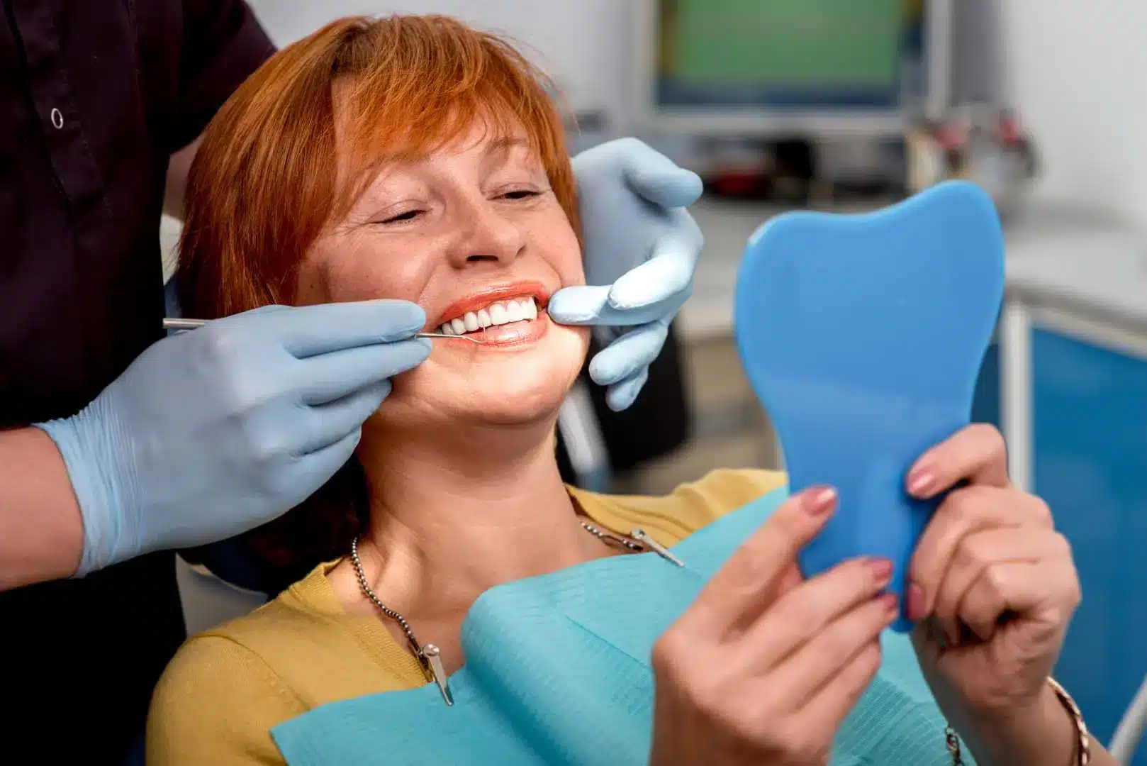 A woman in a dental chair looks happily into a hand mirror at her new smile after a successful CEREC restoration, while a dental professional works on her mouth.
