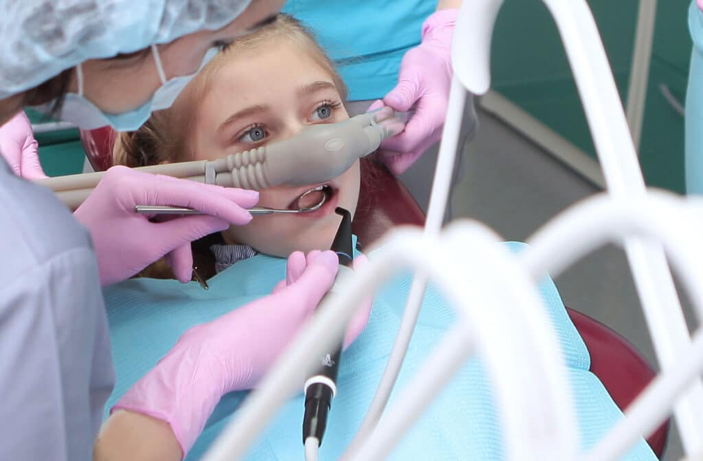 A young female patient receiving nitrous oxide or laughing gas through a nasal mask while two dental professionals, wearing pink gloves, work on her teeth with tools.